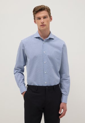 Uomo che indossa una camicia a quadretti piccoli azzurro chiaro, infilata in pantaloni neri, in piedi con una mano in tasca davanti a uno sfondo uniforme.