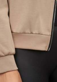 Beige zip-up jack met ribbels aan de onderkant en mouwen, gemaakt van gladde stof. Heeft een zilveren rits aan de zijkant tegen een zwarte achtergrond.