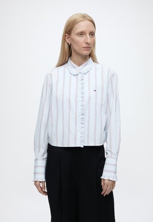 Frau mit glatten blonden Haaren, die ein hellblau gestreiftes Langarmshirt mit Rüschen und schwarze Hosen trägt, steht vor einem schlichten Hintergrund.