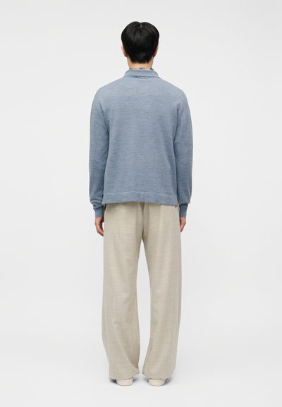 Άνδρας που στέκεται, γυρισμένος με την πλάτη, φορώντας ανοιχτό μπλε μπλούζα με μακριά μανίκια, μπεζ παντελόνι με φαρδιά πόδια και άσπρα παπούτσια, σε απλό λευκό φόντο.