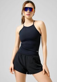 Sort sports tank top med tynde stropper og t�ætsiddende pasform, parret med løse sorte shorts. Modellen bærer refleksionssolbriller.
