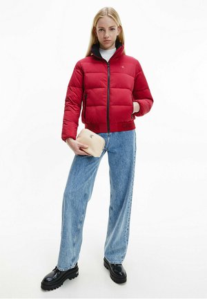 Giacca a vento rossa con zip e colletto neri, con un logo sull'elastico in vita; abbinata a jeans a gamba larga blu chiaro e stivali neri.