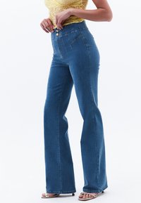 Jeans de mezclilla azul de cintura alta con pierna acampanada. Cuentan con un cierre de botones en la parte delantera y detalles de costura vertical. Textura suave y diseño casual.