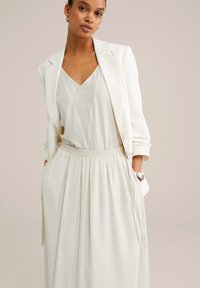 Blazer bianco su un morbido vestito color crema chiaro con scollatura a V. Il vestito presenta una vita elasticizzata arricciata e tasche laterali.