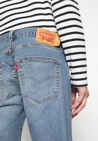 Vaqueros de mezclilla azul claro con un parche de cuero marrón, cinco bolsillos y una etiqueta de logo roja en la parte trasera. Camiseta a rayas en blanco y negro visible.