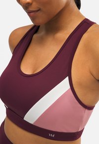 Sujetador deportivo burdeos con un patrón diagonal en blanco y rosa claro. Presenta un escote redondeado y un diseño ajustado para brindar soporte. Tela texturizada.