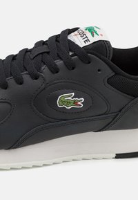 Baskets en cuir noir avec une tige texturée, dotées de lacets noirs, d'une semelle intermédiaire blanche et d'un logo crocodile vert brodé sur le côté.