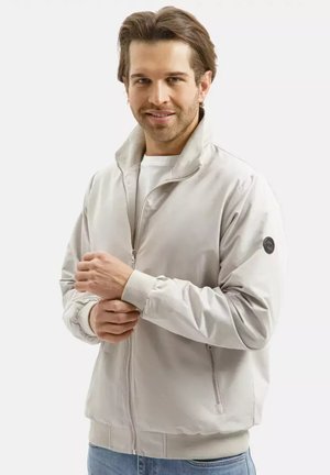 Mężczyzna z jasnobrązowymi włosami w jasnobeżowej kurtce na zamek, poprawiający rękaw, uśmiechający się na jednolitym białym tle.