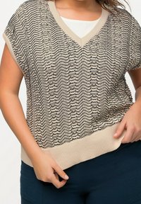 Beige stickad tröja med V-ringning, med svarta zigzag-mönster, korta ärmar och ribbad fåll. Matchad med mörka byxor.