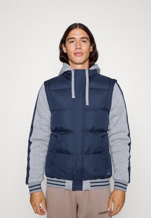 Navy puffer vest with gray hoodie sleeves, kangaroo pockets, ribbed hem, and gray drawstrings; features quilted texture and sporty design.