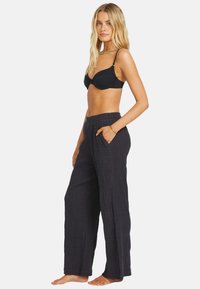 Pantalones anchos negros hechos de tejido texturizado, con una cinturilla alta y bolsillos laterales. La modelo posa descalza, mostrando el ajuste de los pantalones.