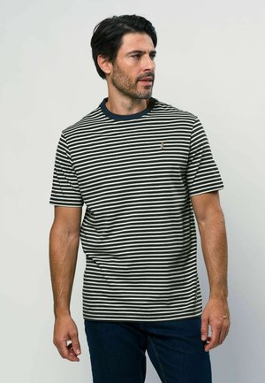 Sötét hajú, szakállas férfi fekete-fehér csíkos pólóban és sötét farmerral, jobb felé néz egy egyszínű háttér előtt.