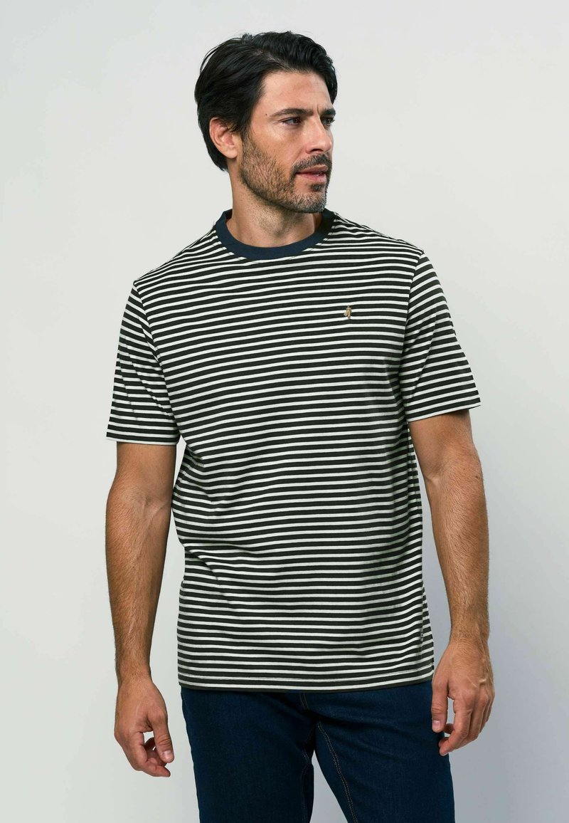 Homme aux cheveux foncés et barbe portant un t-shirt rayé noir et blanc et un jean foncé, regardant vers sa droite sur un fond uni.