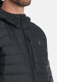 Schwarze gepolsterte Jacke mit hohem Kragen, Frontreißverschluss und einem Spinnenlogo. Verfügt über eine horizontale Steppstruktur und glattes Material.