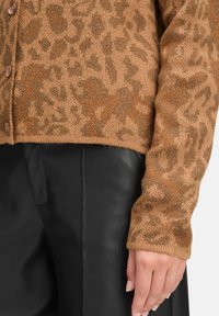 Cardigan en imprimé léopard marron avec un tricot doux et texturé et un devant boutonné. Porté avec un pantalon en cuir noir, mettant en valeur les matériaux contrastés.