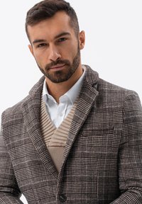 Ruskean ruudullinen bleiseri, jossa on shaalikaulus, ja sen alla on teksturoidusta beigeestä valmistettu liivi valkoisen paidan päällä, tummien nappien ja sulavan muotoilun korostamana.