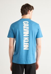 T-shirt in cotone blu con maniche corte, caratterizzata da grande testo bianco "CALVIN KLEIN" stampato verticalmente sul retro. Tessuto liscio, vestibilità classica.