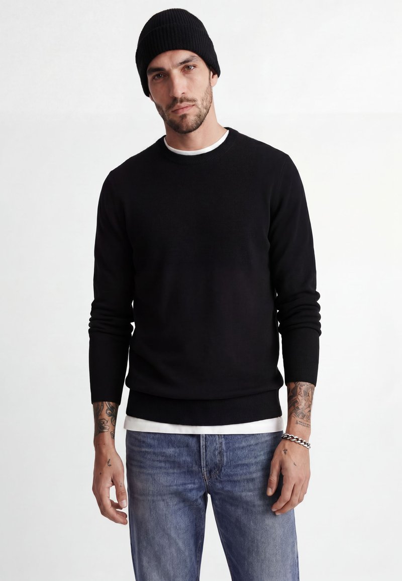 Homme portant un bonnet noir, un pull noir par-dessus une chemise blanche, un jean bleu, avec des tatouages sur les bras et un bracelet chaîne au poignet droit.