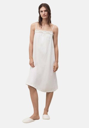 Vrouw staand, gekleed in een mouwloos wit nachthemd met kanten details en witte slippers, tegen een effen lichte achtergrond.