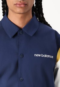 Chemise boutonnée marine avec col, arborant le logo "new balance" en broderie blanche, fabriquée en tissu lisse avec des manches de couleur contrastante.