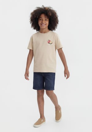 Enfant souriant aux cheveux bouclés marchant, portant un T-shirt beige avec un motif de fruits, un short en jean foncé et des chaussures beiges à enfiler sur fond blanc.