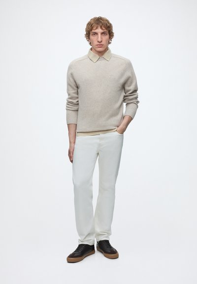 Homme portant un pull beige avec col, pantalon blanc et baskets noires, debout avec une main dans la poche devant un fond blanc.