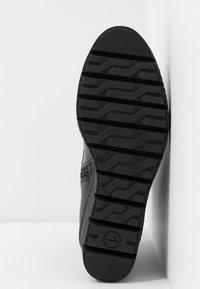 Semelle de chaussure noire avec des motifs de profondeur et une taille marquée "37", positionnée verticalement sur un fond blanc dans un coin.