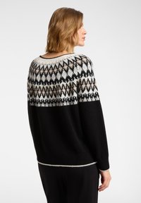 Kobieta zwrócona bokiem, ubrana w czarny sweter z geometrycznym wzorem na karczku w kolorach białym, beżowym i czarnym, na jednolitym jasnym tle.