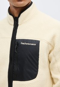 Άνδρας που φοράει μπεζ φλις μπουφάν με μαύρη τσέπη στο στήθος με φερμουάρ, κεντημένη με το λογότυπο «Peak Performance».