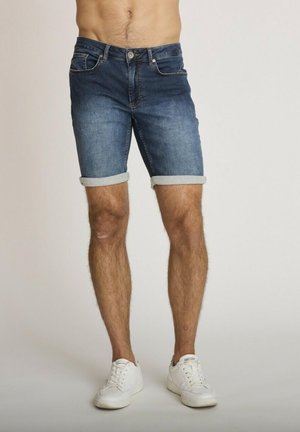 Homme portant un short en jean bleu foncé retroussé et des baskets blanches, debout devant un fond clair uni.