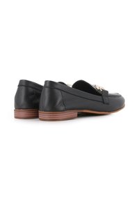Mocasines de cuero negro con una suela plana marrón, que cuentan con un acento metálico decorativo en la parte superior. Textura suave con detalles de costura mínimos.