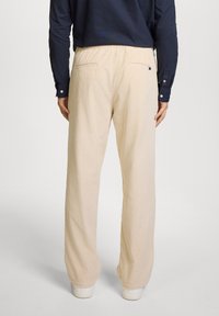Pantalones de pana en beige claro con una superficie texturizada y acanalada. Cuentan con una cintura elástica y dos bolsillos traseros. Se llevan con una camisa de manga larga azul marino.