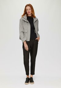 Graue, taillierte Jacke mit hohem Kragen, zwei Fronttaschen, schwarzer Ripp-Pullover, schwarze schmal geschnittene Hose und schwarze Sneakers mit beigen Sohlen.