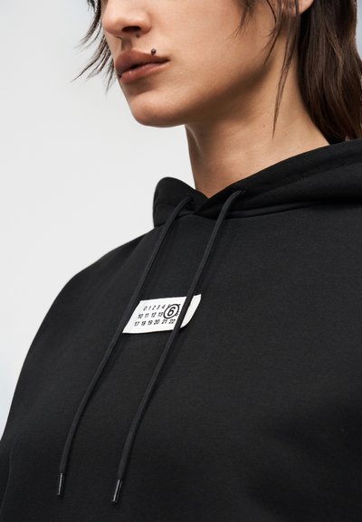 Κοντινό πλάνο ενός ατόμου που φοράει μαύρη μπλούζα με κουκούλα, με μια λευκή ετικέτα που έχει αριθμούς και έναν κύκλο γύρω από τον αριθμό 6 στην περιοχή του στήθους.