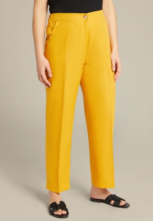 Pantaloni gialli su misura con una texture liscia, dotati di chiusura con bottone e tasche laterali. Abbinati a sandali neri piatti.
