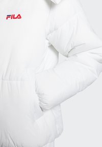 Hvid dunjakke med lynlåslukning foran, synlig lomme og FILA-logo i rød og blå. Har en blød, polstret tekstur for isolering.