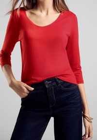 Rotes Langarmoberteil mit Rundhalsausschnitt, kombiniert mit dunklen Jeans. Das Oberteil hat eine glatte Textur, während die Jeans eine klassische Passform haben.