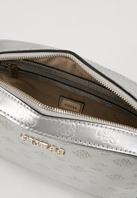 Pochette sintetica argentata con chiusura a zip. Interno con fodera beige e etichetta con hardware dorato. Dettaglio logo testurizzato sull'esterno.