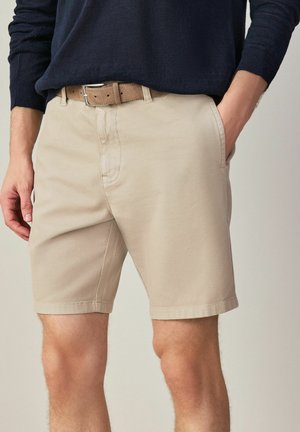 Mann trägt beige Shorts mit seitlichen Taschen und einem braunen Gürtel, kombiniert mit einem dunklen Langarmhemd, die Hände entspannt an den Seiten.