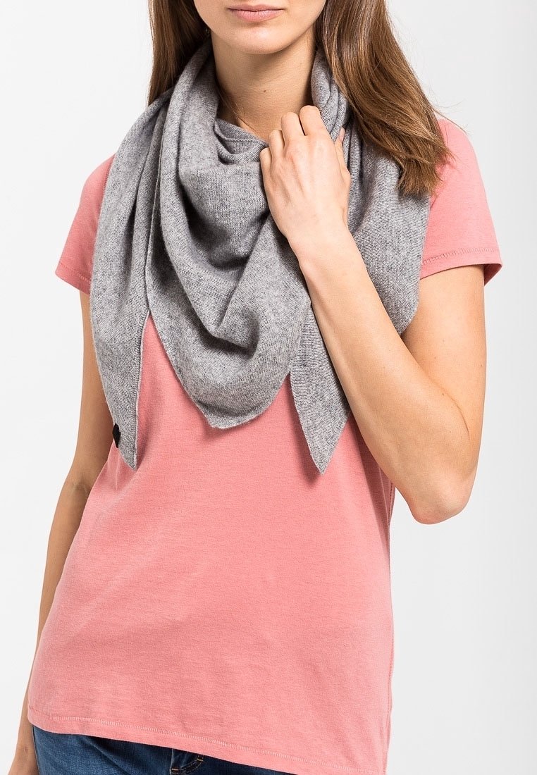 Femme portant un t-shirt rose à manches courtes et une grande écharpe grise drapée sur les épaules, tenant l'écharpe près du cou avec la main droite.