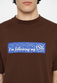 Brown t-shirt featuring a blue rectangular graphic with white text: "I'M NOT LOST, I'm following my Star Sign," accented by small stars.