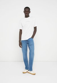 Λευκό βαμβακερό μπλουζάκι με τυπωμένο κείμενο, σε συνδυασμό με ανοιχτό μπλε τζιν παντελόνι και κρεμ αθλητικά παπούτσια με ταμπά σόλα. Το μοντέλο στέκεται μπροστά σε λευκό φόντο.