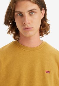 Mosterdgele sweatshirt met een ronde hals, voorzien van een klein rood Levi's-logo op de borst. De stof lijkt zacht met een gestructureerde afwerking.