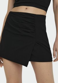 Mini-jupe noire avec texture froncée sur un côté, ourlet asymétrique et finition en tissu lisse. Convient pour plusieurs options de style.