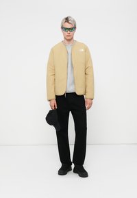 Beige polstret jakke med glidelås, grå genser, svarte bukser, svarte joggesko og svart caps. Solbrillene har grønne og blå detaljer.