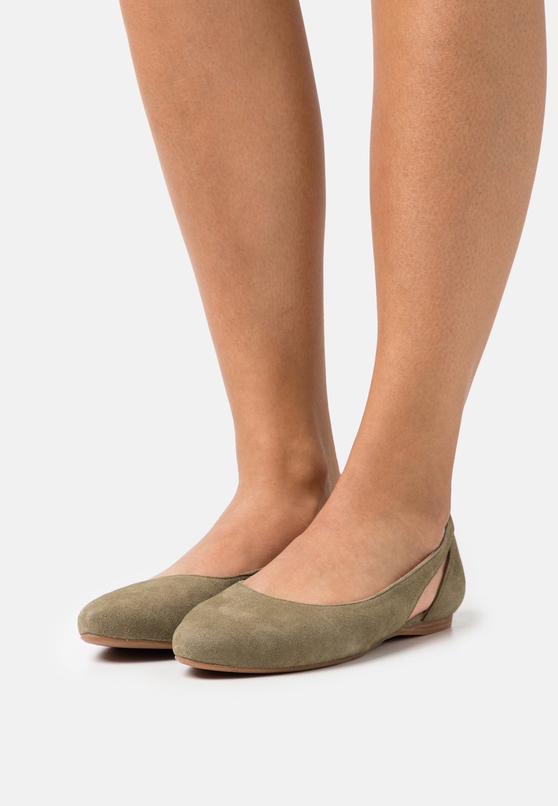 Pieds portant des ballerines en daim vert olive avec un petit talon et un détail découpé près du talon.