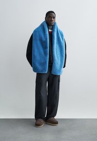 Νεαρός άνδρας όρθιος, φοράει μια μεγάλη μπλε χνουδωτή κασκόλ, μαύρο μπουφάν, σκούρο παντελόνι cargo και καφέ μοκασίνια με σχέδια, με γκρι φόντο.