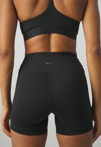 Femme portant un brassière de sport noire Nike et un short de sport noir taille haute, vue de dos sur un fond gris uni.