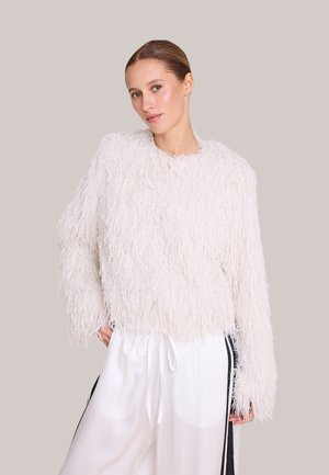 Kvinne med håret greidd bakover, iført en hvit, strukturert, lodden genser med lange ermer og løse hvite bukser med svarte striper.