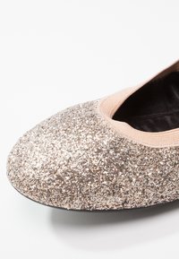 Gros plan sur une ballerine couverte de paillettes, avec une bordure élastique beige et une semelle intérieure noire sur fond blanc.